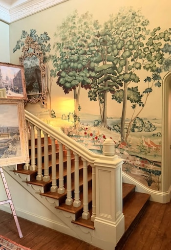 Amplia escalera de madera con barandilla blanca crema en un vestíbulo con un mural que representa árboles, un paisaje campestre y un espejo barroco