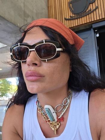 Emilia Attias lleva anteojos de sol, una pañoleta naranja en el cabello, una camiseta blanca sin mangas y varios collares