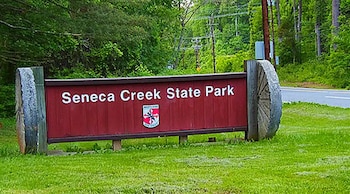 Un letrero de madera y piedra dice 'Seneca Creek State Park' con un emblema de Maryland. Pasto, árboles, postes de luz y una carretera al fondo