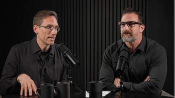 Dos hombres con gafas y camisas oscuras se encuentran frente a micrófonos de mesa en un estudio de fondo negro