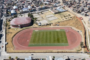Vista aérea de una pista de atletismo con césped central, un coliseo de techo rojo, canchas auxiliares y viviendas urbanas alrededor