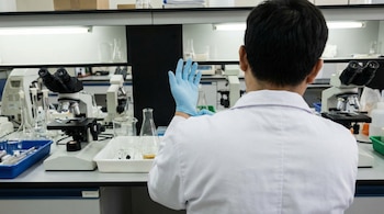Científico de espaldas en laboratorio poniéndose un guante celeste. Microscopios y material de vidrio de laboratorio son visibles en el fondo.