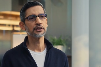 Perto de Sundar Pichai, um homem de meia-idade, de óculos, barba e cabelos escuros, vestindo uma jaqueta azul marinho com gola branca.