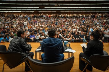 Tres personas sentadas de espaldas en sillones sobre un escenario frente a una multitud en las gradas de un auditorio techado