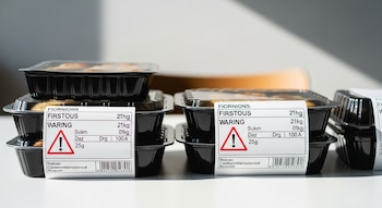 Envases de plástico negros apilados en una mesa blanca con etiquetas que muestran texto ficticio y un triángulo de advertencia rojo.