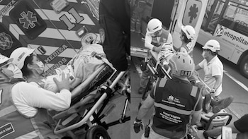 Varias personas, incluyendo personal de emergencia con uniformes y cascos, atienden a dos heridos tendidos en camillas, junto a una ambulancia y un bus, en blanco y negro
