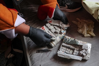 Manos con guantes negros abren paquetes envueltos en periódico revelando su contenido, sobre un colchón. Una persona con un disfraz naranja y rojo está cerca