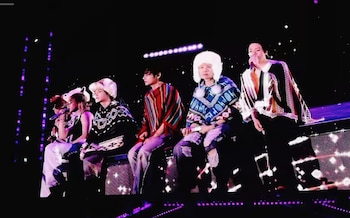 Integrantes de BTS sentados y de pie en un escenario oscuro con ponchos de lana estampados, gorros blancos y micrófonos
