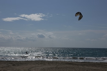Vista panorámica de una playa con un kitesurfista sobre las olas del mar, bajo un cielo parcialmente nublado. Un cometa de kitesurf flota en el aire