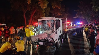 Vehículo Isuzu blanco con frontal dañado, rastra sin carga, rescatistas con uniformes amarillos, policías, personas en la carretera, luces de emergencia.