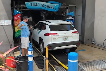 Un hombre con gorra y camiseta azul limpia un SUV blanco en un lavadero automático, utilizando un cepillo largo mientras el vehículo avanza por el túnel de lavado