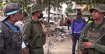 Dos militares con uniforme verde y sombreros, un civil con gorra y mascarilla, otro civil de camisa azul. Escombros, retroexcavadora y personas al fondo