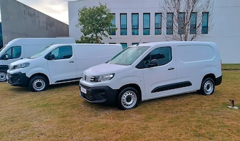 Varias vans blancas Peugeot Partner y Citroën Berlingo estacionadas en un césped frente a un edificio moderno con ventanas rectangulares y un árbol
