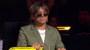 Un hombre con cabello castaño, anteojos de sol, blazer verde oliva y una cadena dorada en el cuello, sentado frente a una superficie amarilla.