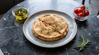 Pizzolo cortado en un plato gris sobre mármol negro, con un tazón de tomates cherry, un tazón de aceite y ramitas de romero.