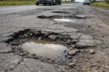 Una carretera de asfalto con múltiples baches llenos de agua. Se observan un camión y varios automóviles a la distancia bajo un cielo nublado.