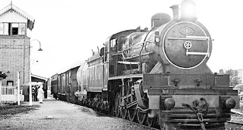 Imagen en blanco y negro de una locomotora a vapor con el número 191 y vagones, parada en una estación de ladrillo con un edificio y algunas personas
