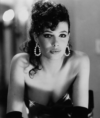 Retrato en blanco y negro de Kelly LeBrock con cabello rizado recogido, grandes pendientes, top sin tirantes y guantes. Mira directamente a la cámara
