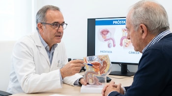 Doctor con bata blanca y gafas señala un modelo anatómico de próstata a un paciente mayor. Detrás, un monitor muestra imágenes de próstata y cáncer.