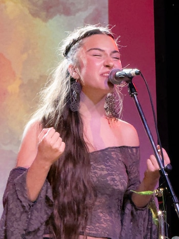 Una persona con cabello largo toca una guitarra acústica con los ojos cerrados frente a un micrófono en un escenario iluminado