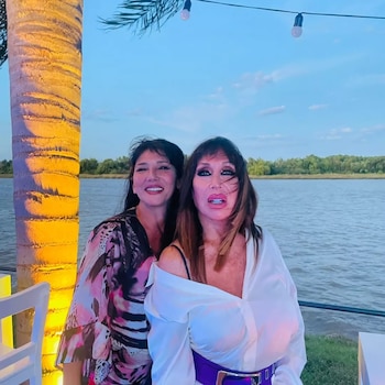 Moria Casán y su amiga Zeta posan junto a una palmera iluminada con un amplio cuerpo de agua detrás