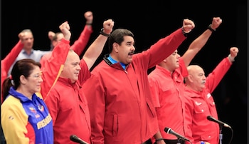 Nicolás Maduro, con chaqueta roja, y otros funcionarios con camisas rojas levantan sus puños derechos en un escenario oscuro, con micrófonos en primer plano