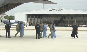 Agentes federales armados y uniformados custodian a personas vestidas con uniformes celestes en la pista de un hangar
