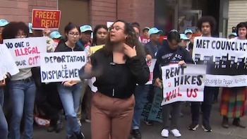 Un grupo de personas, incluyendo jóvenes, participa en una manifestación callejera, sosteniendo pancartas con mensajes en inglés sobre alquileres, deportación e inmigración