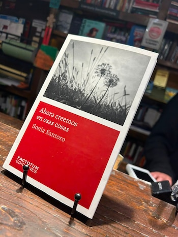 Libro con cubierta roja y superior blanca con foto en blanco y negro de dos dientes de león, título 'Ahora creemos en esas cosas' de Sonia Santoro. Exhibido sobre madera