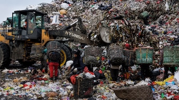 Un vertedero lleno de basura con un cargador frontal amarillo de Hyundai. Varias personas con cestas en la espalda y el suelo cubierto de residuos
