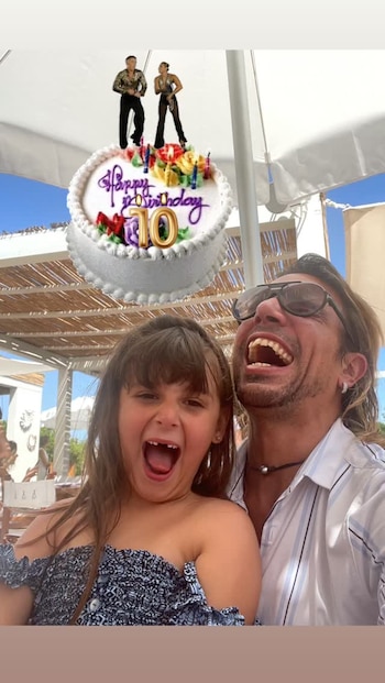 Un hombre con gafas de sol y una niña sonríen a la cámara bajo un toldo blanco, con un dibujo digital de un pastel de cumpleaños superpuesto