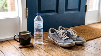 Una taza de café sobre un posavasos, una botella de agua y un par de zapatillas sobre un felpudo ante una puerta azul.