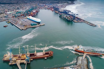Vista aérea de un puerto comercial con numerosos buques de carga, incluyendo un portacoches y portacontenedores, y grúas en muelles concurridos. Se ve una ciudad en el fondo y el mar