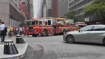 Camión de bomberos rojo y blanco, coche sedán plateado, personas caminando, edificios altos, varias banderas de Estados Unidos, dos bolardos