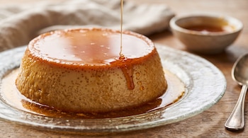 Un flan sobre un plato de vidrio transparente recibe caramelo líquido en una mesa de madera con una cuchara al lado.