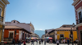 Vista de una calle concurrida en Guatemala, bordeada por edificios coloniales de colores vibrantes y puestos de venta con sombrillas. Personas caminan bajo un cielo azul claro.