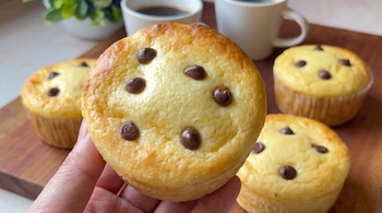 Mano que sostiene un pan de leche circular con chispas de chocolate, con tres panes más y dos tazas blancas de fondo.