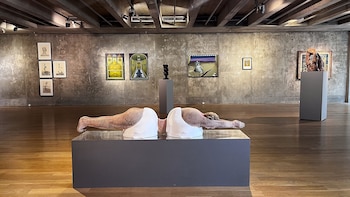 Interior de una sala de exposición de arte con una escultura central de un cuerpo fragmentado sobre un pedestal, rodeada de pinturas y esculturas en paredes de hormigón