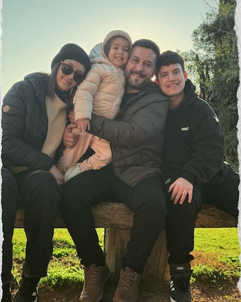 Cuatro personas, dos adultos y dos jóvenes, posan sonrientes en un banco de madera. El fondo muestra vegetación y un cielo despejado al atardecer