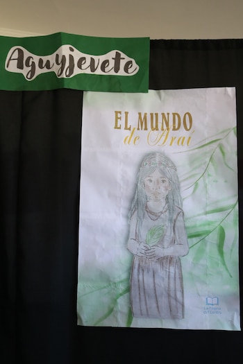 Cartel verde con la palabra Aguyjevete colocado sobre un fondo negro junto a una lámina ilustrada de una niña con el texto El Mundo de Araí