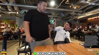 Dos hombres en un restaurante: uno de pie con camiseta negra y el otro sentado de blanco gesticulando. Hay otras personas comiendo en mesas al fondo