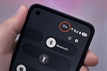 Primer plano de mano sosteniendo teléfono móvil. Pantalla oscura muestra ícono de llave en círculo naranja, señales de red y Bluetooth.