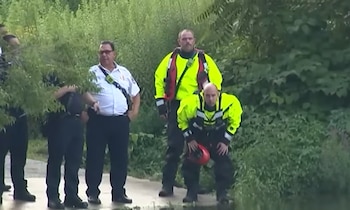 Cinco hombres, algunos con uniforme y otros con trajes de rescate amarillos, están en la orilla de un río. Uno de los hombres se agacha con un casco rojo