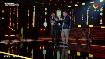Dos hombres en un escenario. Uno con traje sujeta un premio dorado. El otro con chaqueta sostiene un micrófono. Fondo con luces y texto del programa