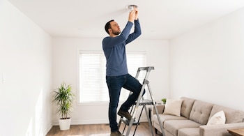 Hombre sobre una escalera metálica ajustando una bombilla blanca en un aplique de techo dentro de una habitación con sofá y planta.