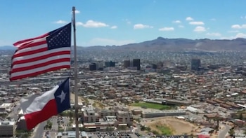 Una vista aérea de la bandera de Estados Unidos y la bandera de Texas ondeando sobre un paisaje urbano extenso con edificios, montañas al fondo y cielo azul