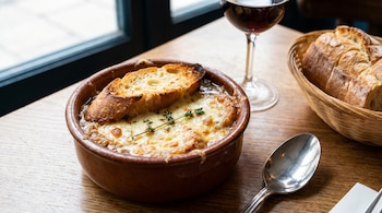 Un tazón de sopa de cebolla a la francesa con queso gratinado y croutones sobre una mesa de madera. Cerca, una copa de vino, una cuchara y pan en una cesta.
