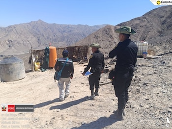 Agentes de policía y un funcionario caminan por un terreno árido de montaña con estructuras rústicas y tanques de agua al fondo