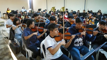 Grupo de jóvenes sentados tocando violines en un ensayo, con atriles y algunos instrumentos de viento metálico en el fondo