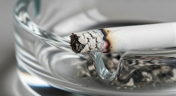 Primer plano de un cigarrillo parcialmente consumido, con el extremo encendido y la ceniza gris, apoyado en un cenicero de vidrio transparente con bordes curvos.